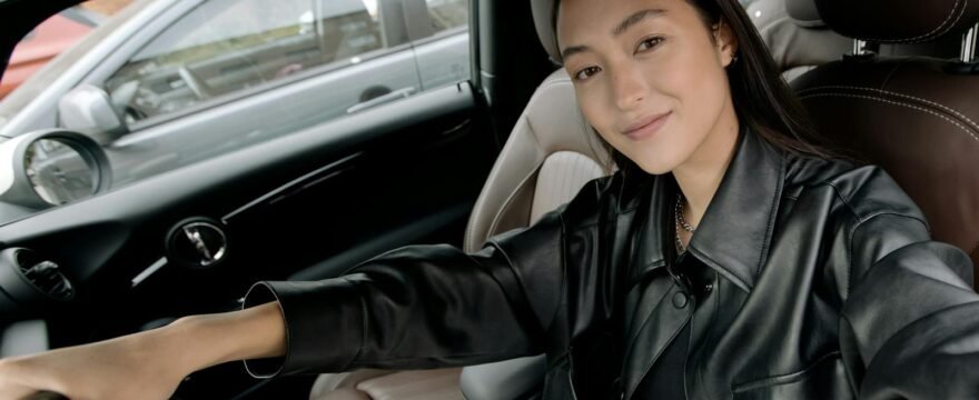 A young woman in a black leather jacket takes a selfie inside a modern car, showcasing a relaxed pose.